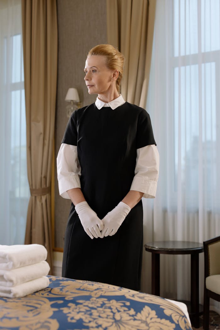 services-03 Chambermaid in uniform standing in hotel room with folded linens, exuding professionalism.