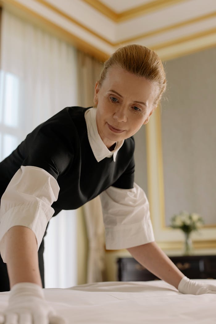 hero-img-01 A housekeeper in uniform skillfully arranges bed linen in a luxurious hotel room.